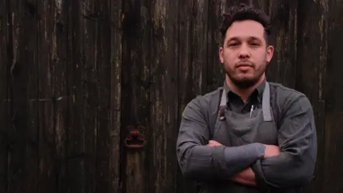 Abbie Morris Corrin Harrison in grey apron and dark grey shirt and arms crossed. He is standing in front of a wooden door and has dair hair and short trimmed dark facial hair