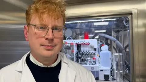 The photo shows a man looking at the camera. He has short, ginger hair and is wearing glasses, a white lab coat and a dark jumper. Behind him is a metal door and a machine with various wires connected to it. 
