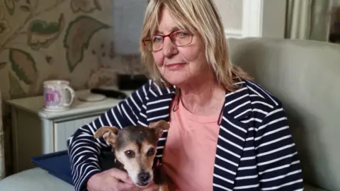 A woman with blonde shoulder-length hair and wearing glasses. She is sitting in an armchair, with her Jack Russell dog on her lap. She is wearing a navy and white striped jacket with a coral coloured T-shirt.