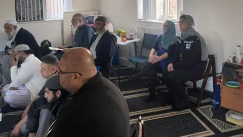 Office of the police, fire and crime commissioner A small group of men sit in a room with white walls.  Some of the men are wearing skull caps. Two women sit at the back - bother are wearing blue scarves over their heads (Danielle Stone and Sophia Parveen)