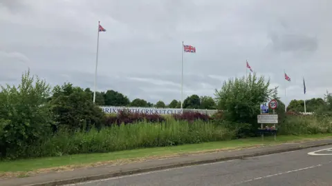 Pete Cooper/BBC In the foreground of the picture is a road. The other side of the road a sign that says "Brixworth Cricket Club" is just visible behind an overgrown hedge. Union flags on tall flag poles are flying in front of a cloudy sky. 