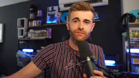 Dean shows off his streaming set-up. He's behind a microphone, posing with a neutral, fairly serious look. He's got a very neat beard and tidy, short hair and wears a t-shirt with thin blue and burgundy stripes. The background is blurred but we can make out gaming memorabilia, including a framed Call of Duty print and several Funko Pop! figures in and out of boxes. Most appear to be Pokemon themed.