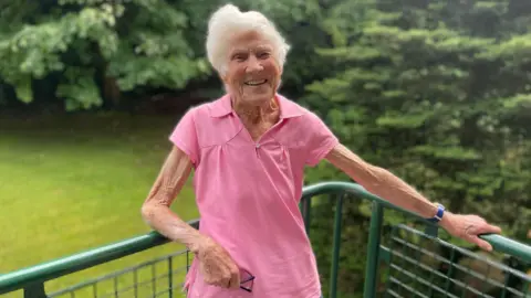 BBC Grace is standing on a balcony, she is wearing a pink t-shirt and smiling at the camera