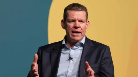 Plaid Cymru Leader Rhun ap Iorwerth speaking at a conference. He is sitting in front of a green and yellow wall. 