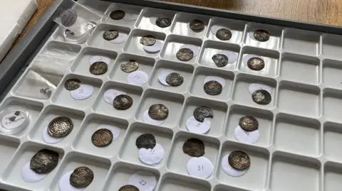 A plastic grey tray containing 30 old silver coins and some fragments of coins. The coins are labelled with small discs of paper. They are small coins, made of silver and struck with a dais.