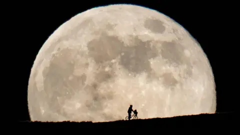 Silhouette of boyish character in an anorak straddling a stationary BMX bike with an object in its front basket on a mountainside framed by the full Moon behind