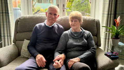 A man and a woman sat next to each other on a sofa holding hands. A plant is to their left and they are sat in front of glass doors.