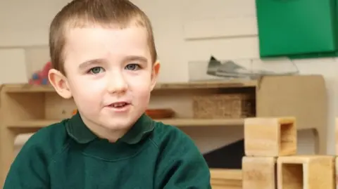 Essex Police Lincoln Button is smiling at the camera, he has blue eyes, short brown hair and is wearing a dark green jumper over a dark green polo shirt. 