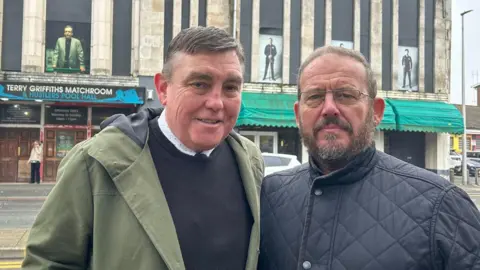 Andrew Morgan is wearing a green coat and dark jumper and Adam Rees has a black winter jacket on. The matchroom has a green awning and a sign saying  Terry Griffiths Matchroom with three metre high picture of Terry playing pool in a competition in the window above