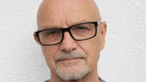 Chris Purkess Chris Purkess stands against a white wall. He is bald with glasses and a goatee beard.