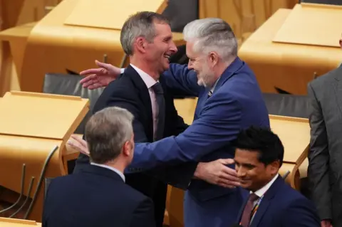 PA Media Liberal Democrat MSP Liam McArthur (left) is congratulated by MSP George Adam in the Scottish Parliament after MSPs approved the assisted dying bill in its first stage