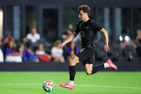 Getty Images Callan Elliot in a black top, shorts and socks in bright pink boots runs forward with the ball at his feet