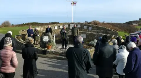 Community members at a previous memorial service at Hammond Memorial. Spectators are standing in front of the memorial while a service is underway. 