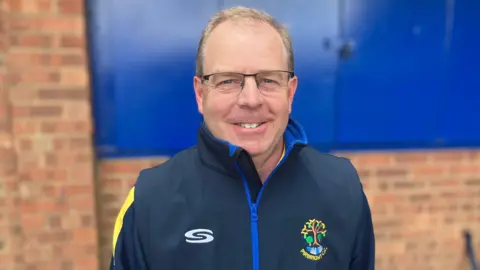 Paul Brennan / BBC A man wearing a dark blue quarter zip top with a logo of a tree on the right side. He is stood in front of a brick building with blue shutters.