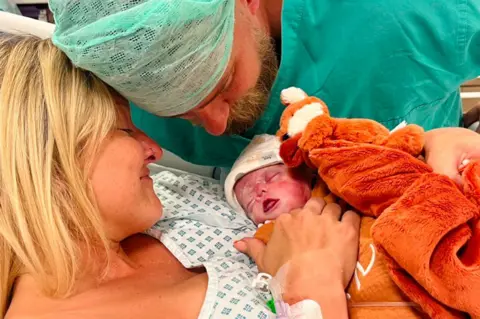 Andrea McCartney Andrea McCartney in a hospital gown holding her newborn baby Kit, with a smile on her face. Her partner is in green surgical scrubs close to his son