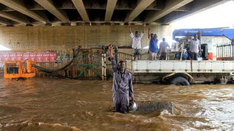 Ebrahim Hamid/AFP/Getty Images A man standing knee deep in dirty water with his hand up in the air. There are other workers behind him who are standing in the back who also have their hands in the air. They appear to be under some sort of bridge. Municipal workers react while operating a pump draining a flooded highway overpass in Sudan's capital Khartoum following heavy rain - Wednesday 27 August 2025