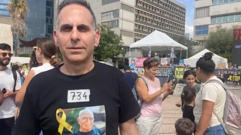 BBC Rotem Cooper stands in a crowded area wearing a black T-shirt bearing the number 734. It has a picture of his father, Amiram Cooper, accompanied by the words "Bring it home now!"