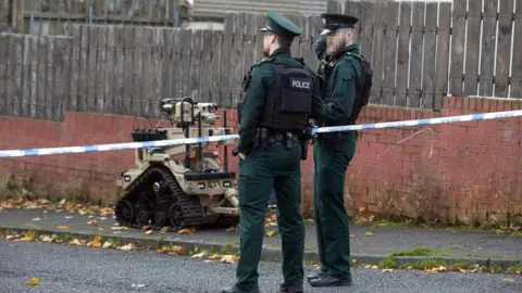 Pacemaker Two male police officers standing at a blue and white police tape. Their faces are blurred and they are wearing a bottle green police uniform with a black bullet proof vest. There is an ATO machine which is a brown colour with black wheels.
