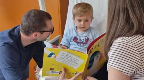 Raffy is sitting on a hospital bed with his dad Phil and mum Sarah. They are reading a book together.