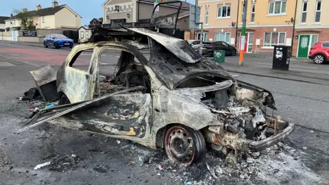 A car is burnt out. Debris and ashes are lying around the road/ A lamppost has been painted green, white and orange.