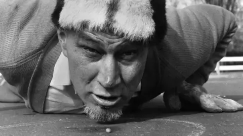 Getty Images A black and white image of Walter Cornelius face on and flat on his stomach with his hands beneath his shoulders. His head is raised and he is looking forward and in front of him on the ground is a pea.