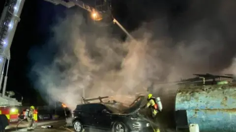 ECFRS Smoke surrounds the wreckage of a car with firefighters dressed in high-vis working around the scene. A large aerial ladder is at one side of the picture and is shooting a jet of water into the smoke.