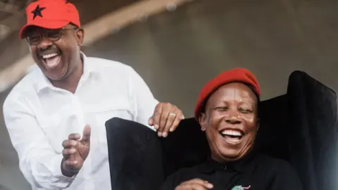 AFP Two men in the picture. One man on the left, Dali Mpofu, wearing a white shirt and a red cap. The man on the right, Julius Malema, is wearing a red cap and a black top.