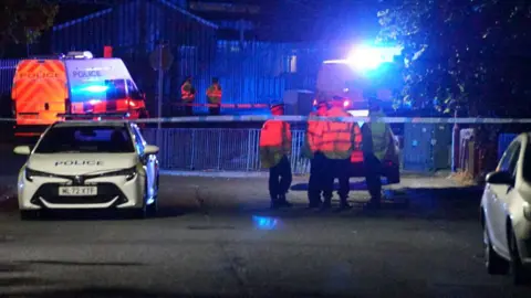 Night time image showing a police cordon with a police car in front. There are four police officers standing close to the cordon.