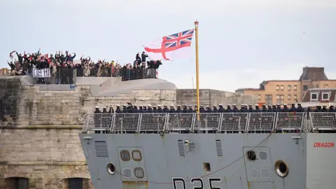 Getty Images Crew members wave goodbye to family and friends as HMS Dragon sets sail from Portsmouth Harbour on March 10