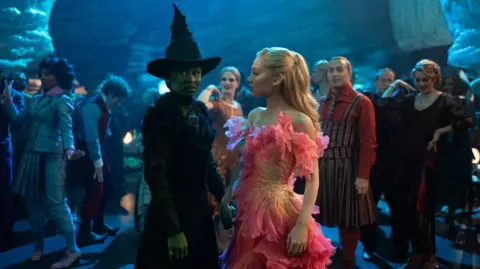 Universal Pictures/Sky Studios Elstree An actor painted green and wearing a withes hat stands with another actor with long blond hair and frilly pink dress, they hold hands, people stand behind them in the background, some are dancing