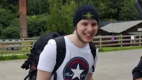 Family Tyran Jones in a family photo. He is smiling. He has a beard. He is wearing a white t-shirt with a converse logo and has a stone island hat on. 