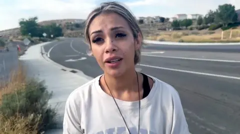 A woman in a white sweatshirt and a necklace standing beside a road.