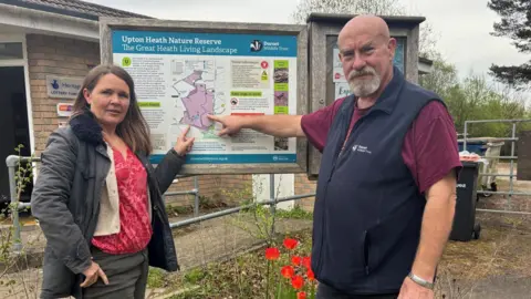 DWT MP Vikki Slade and trust CEO Brian Bleese stand either side a sign displaying a map of Upton Heath and both point at the section which has been bought by the charity. Bleese has a grey goatee beard and is wearing a purple t-shirt with a navy blue gilet. Slade has long brown hair and is wearing grey trousers and a padded jacket with a pink and red floral top underneath.