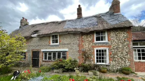 Blake Cottage Trust A flint cottage with a thatched roof, which looks old, damaged, and dilapidated. 