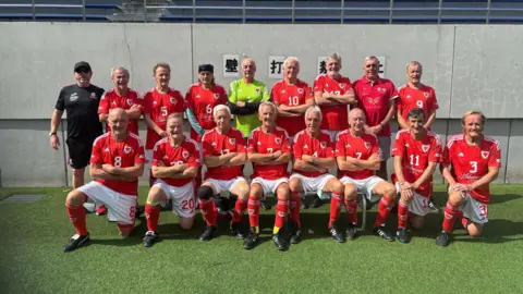 The Wales over-70's team pose for a team photo. They are all wearing full Welsh kits and are in two rows with the people in front kneeling the the people behind stood up.