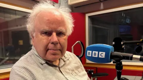 Prof Brian Ripley has wispy white hair and is wearing a grey and white shirt. He is sat at a radio desk with a blue BBC mic in front of him.