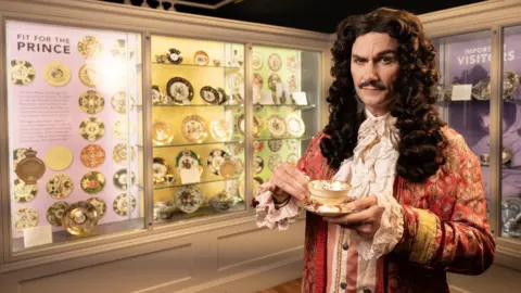A man wearing a long curly dark brown wig, wearing 17th century clothing which consists of a red and gold coat with white frills on the sleeves, and white frills down his chest and hear his neck. He is holding a china cup and saucer, and is standing in front of a museum display of china plates