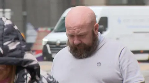 Alan Keelan arriving at court wearing a light grey t shirt. He has a bald head and a light brown beard.