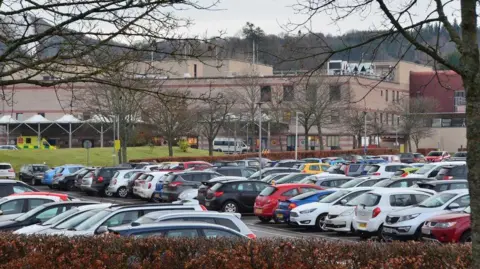 Jim Barton A long pink-coloured building with a packed car park and trees in the foreground.