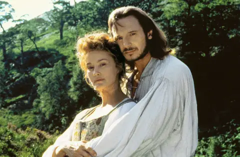 Alamy Liam Neeson and Jessica Lange in period costume in Rob Roy. He embraces her from behind and both look to the camera.