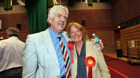 PA Media Rhodri and Julie Morgan stand arm in arm smiling at an election night count in a hall. They both wear red Labour rosettes. He wears a cream linen blazer and blue shirt, she also wears a beige linen jacket and a navy top. 