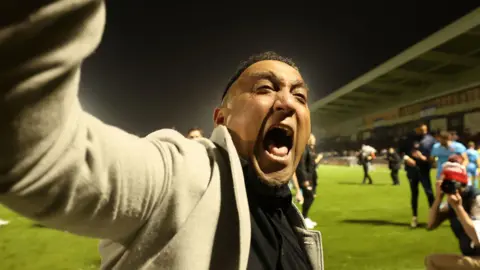 PA George Akhtar shouting in celebration after Kettering Town's win over Northampton. He is on the pitch standing in front of away fans who are out of view. Akhtar has his right arm lifted high in celebration.