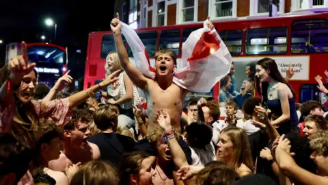 PA A shirtless fan waves a St George's Cross flag while being carried on the shoulders of the crowd in a street in Fulham, south-west London. A red double-decker bus is stopped in the background.