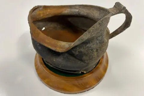 A slightly crumpled metal chamber pot with some signs of rust sitting on a wooden disk with a felt top.