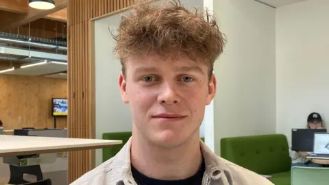 A young man with brown, short frizzly hair is smiling at the camera. He is wearing a white jacket and a black top, and is sitting inside a student learning area.