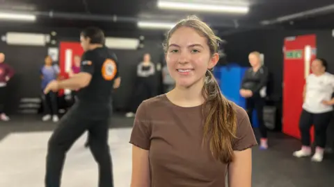 Lucy smiles at the camera as behind her the instructor is showing a demonstration to the class. She is wearing a brown t-shirt and has brown eyes and long brown hair which is tied into a pony tail.