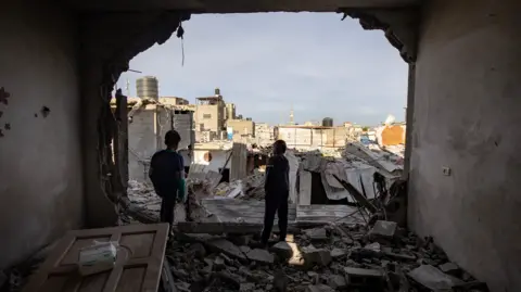 Two Palestinian children stand inside a destroyed apartment, looking out of the space where a wall used to be across a landscape of destroyed buildings