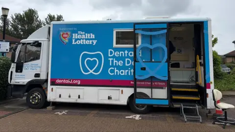 BBC/GRACE STEWART-PIERCY A wide shot of the hub, which is a small lorry with the charity's branding on it. A glass door opens to a set of stairs with a railing, leading into the dental surgery inside.