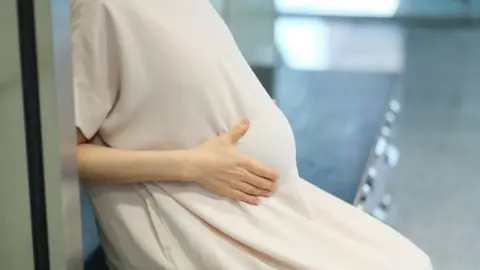 Close-up of a pregnant woman's torso, with her hands cupping her baby bump. She is wearing a long beige dress and sitting on a bench.