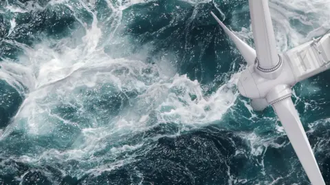 An aerial view of an offshore wind turbine. Visible are the three large white blades and the hub. Below the turbine the sea is green and the water is churned up and there is white froth on the surface of the sea.